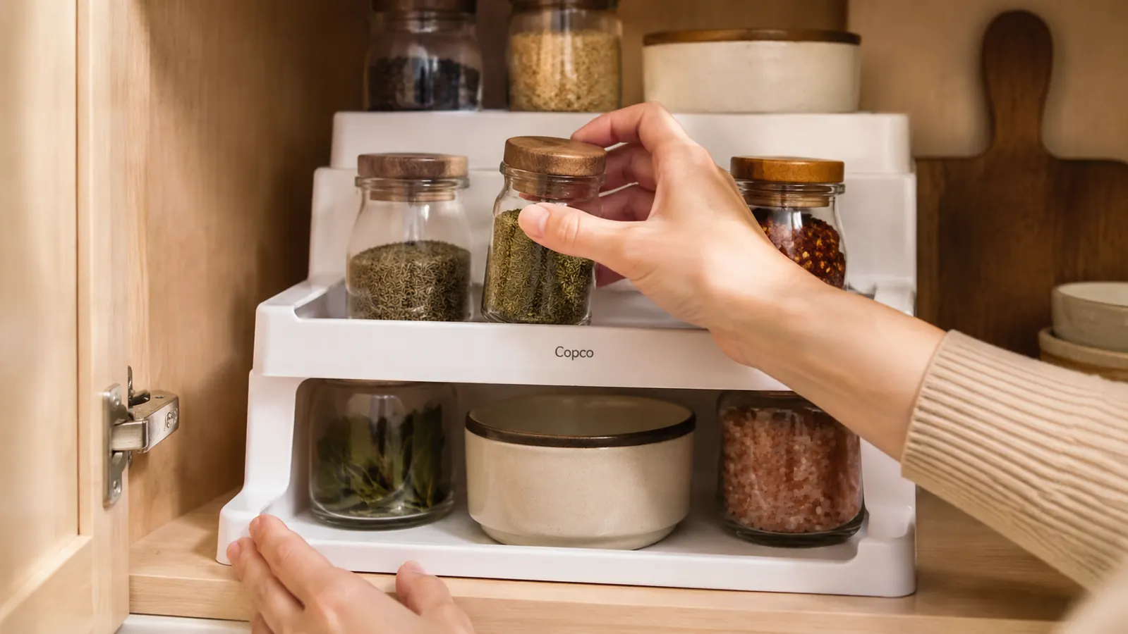 Close-up of a hand placing a spice jar onto the second tier of a Copco spice rack inside a cabinet
