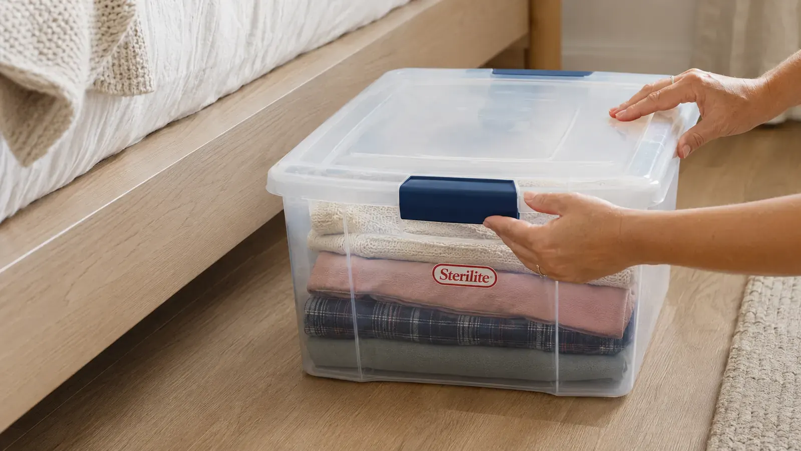 Clear Sterilite latching bin with lid closed, visible folded clothes inside, sitting on a bedroom floor next to a bed
