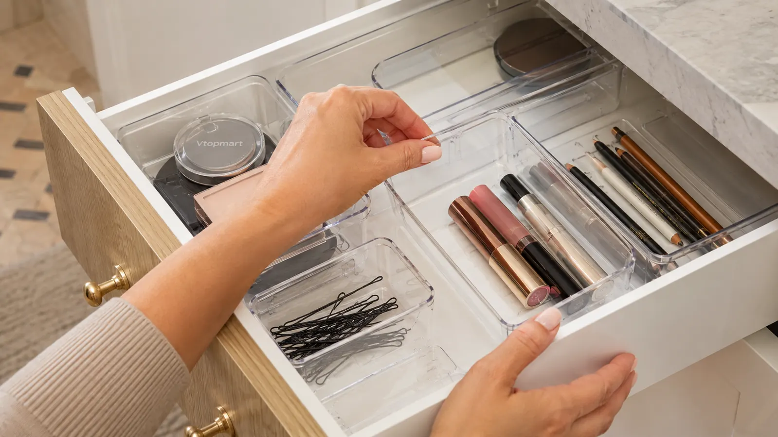 Hands placing small clear plastic organizer bins into a bathroom vanity drawer with makeup and hair accessories