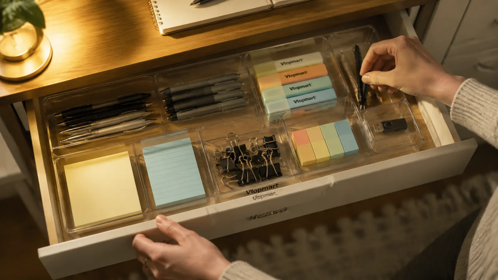 Home office desk drawer organized with clear plastic bins holding pens, sticky notes, paper clips, and USB cables